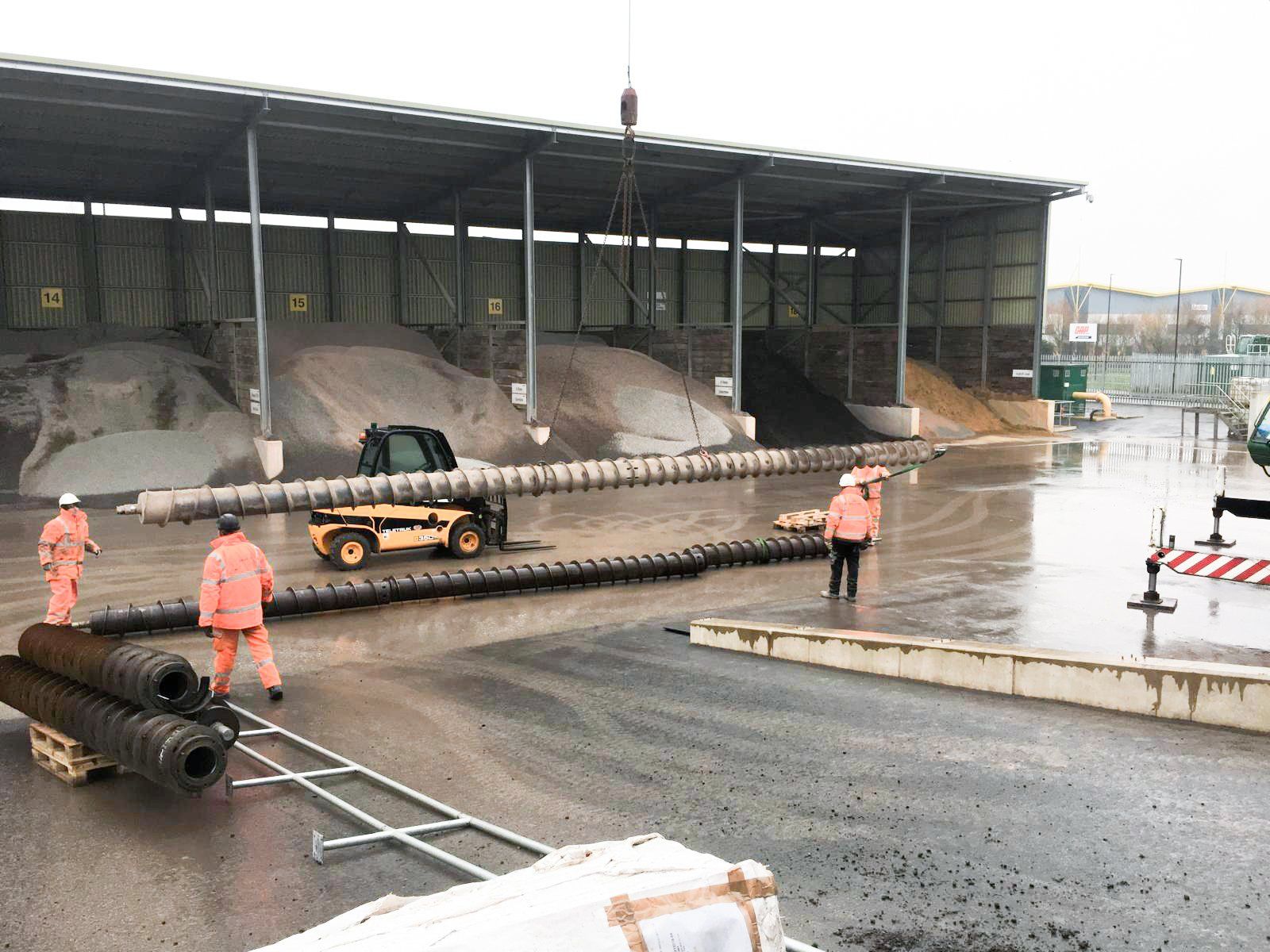 Construction workers in orange safety gear and helmets working outdoors, installing a large pipe on a wet, paved surface near a storage shed with sand and gravel piles in the background.