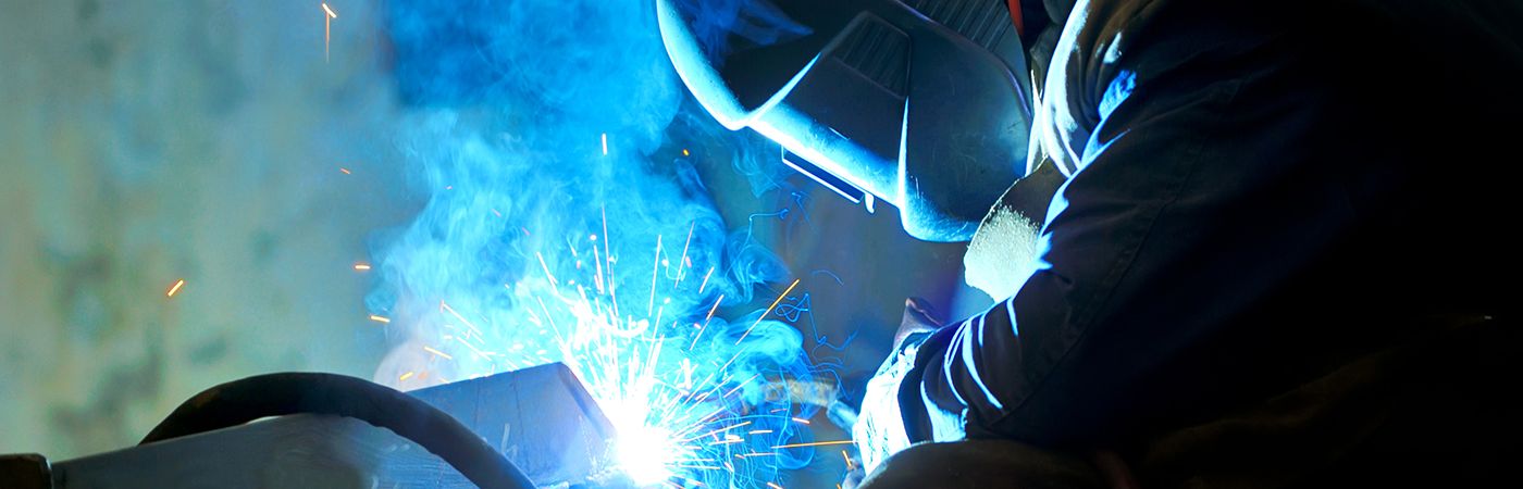A person welding metal while wearing protective gear, including a welding helmet and gloves, with bright sparks and blue light from the welding process.