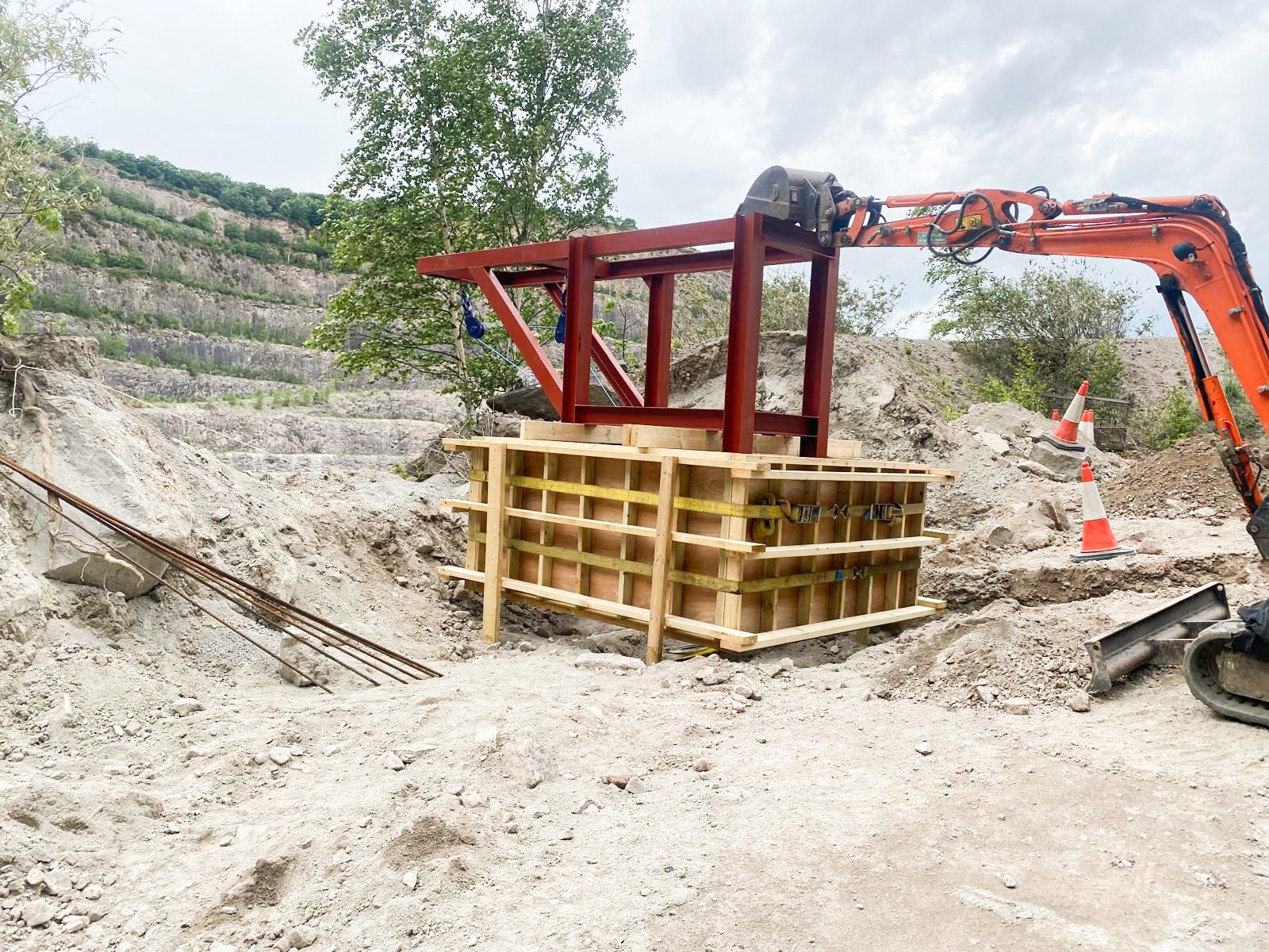 Construction site with a ladder frame and wooden support structure, orange excavator, and traffic cones on a dirt and rocky hillside.